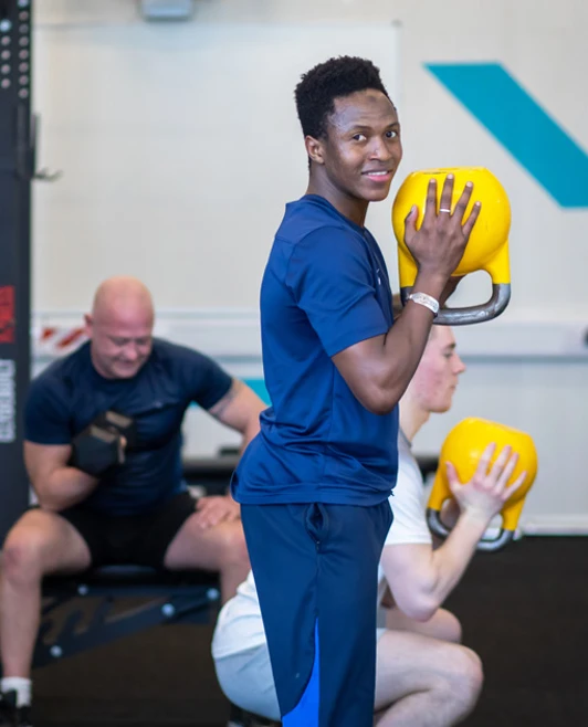 Student smiling while holding a yellow kettlebell during a strength training session at the gym. Student smiling while holding a yellow kettlebell during a strength training session at the gym.